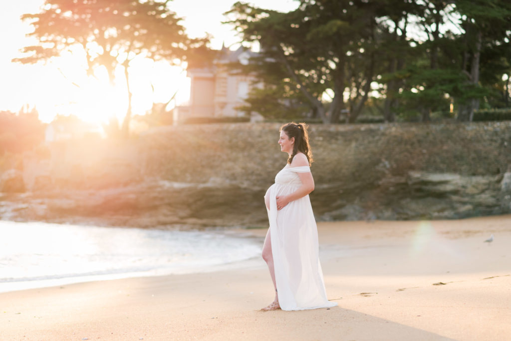Femme enceinte sur la plage en robe blanche en voile au coucher du soleil