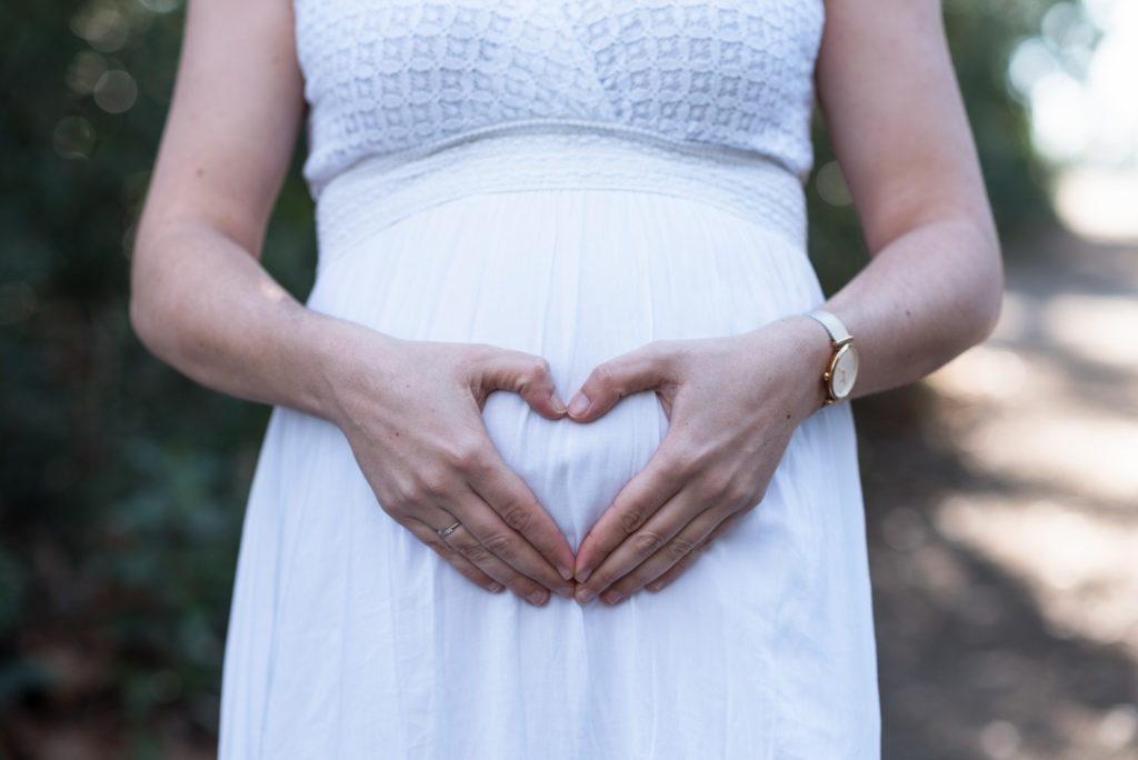 Future maman en robe blanche faisant un cœur sur son ventre avec ses mains